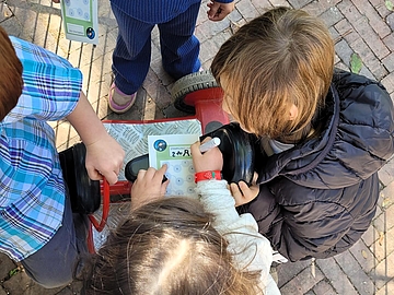 Drei Kinder sitzen eng beisammen, eines davon auf einem roten Kinderfahrzeug. Sie halten Stempelpässe in den Händen und stempeln konzentriert Felder aus. Die Szene findet draußen auf einem gepflasterten Untergrund statt.