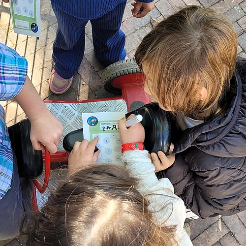 Drei Kinder sitzen eng beisammen, eines davon auf einem roten Kinderfahrzeug. Sie halten Stempelpässe in den Händen und stempeln konzentriert Felder aus. Die Szene findet draußen auf einem gepflasterten Untergrund statt.