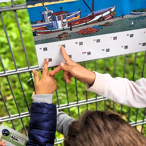 Mehrere Kinder zeigen auf ein farbig illustriertes Plakat, das an einem Zaun befestigt ist. Unten sind QR-Codes mit Sprachkürzeln wie „AR“ für Arabisch und „DE“ für Deutsch abgebildet. Ein Kind hält einen Stempelpass in der Hand. Im Hintergrund ist grüne Wiese zu sehen.