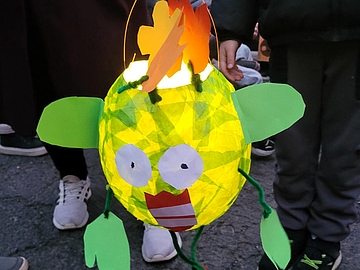 Auf dem Bild ist eine leuchtende, runde Laterne in kräftigem Gelbgrün zu sehen. Sie hat große, weiße Augen, einen roten Mund und grüne Flossen aus Papier. Oben auf der Laterne sind orangefarbene Papierflammen befestigt. Die Laterne wird von einem Kind an einem Laternenstab getragen. Im Hintergrund sieht man weitere Kinder und Erwachsene beim Laternenumzug.
