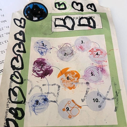 Ein Stempelkärtchen mit zehn durchnummerierten Kreisen liegt auf einem Tisch. Acht Felder sind bunt gestempelt – mit Fischmotiven in verschiedenen Farben. Der Pass ist oben und an der Seite mit schwarzen Herzen bemalt, die mit Filzstift gezeichnet wurden. Ein Kästchen für den Namen ist noch leer. Im Hintergrund ist ein Kalenderblatt mit Datumsangaben zu sehen.