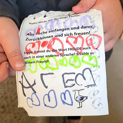 Ein Kind hält einen gestalteten Papierzettel in der Hand. Auf dem Zettel steht: „Alle Fische einfangen und dann: Zurücklehnen und sich freuen!“ sowie die Frage: „Kennst du das Wort FREUDE auch noch in einer anderen Sprache?“ Der Zettel ist bunt mit Herzen, Buchstaben und einem Vogel bemalt.
